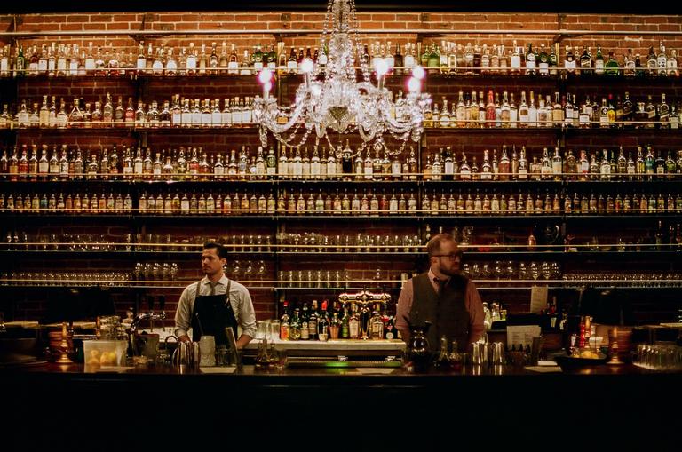 Two men stand behind a bar with many bottles of alcohol on shelves and a chandelier hanging above them.