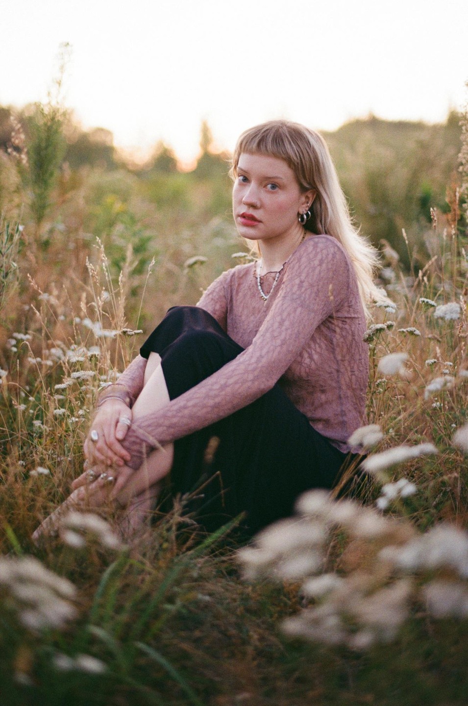 A woman with blonde hair and bangs sits in a field of tall grass and wildflowers at golden hour.