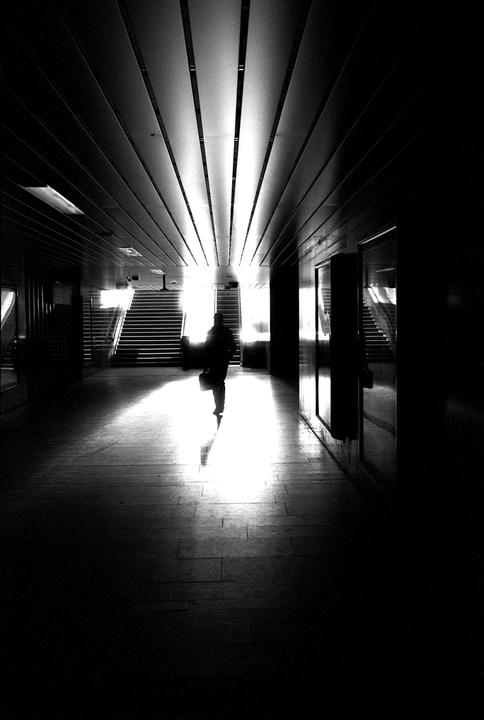 A person walks through a dark hallway towards a bright light at the end, with stairs visible on either side.