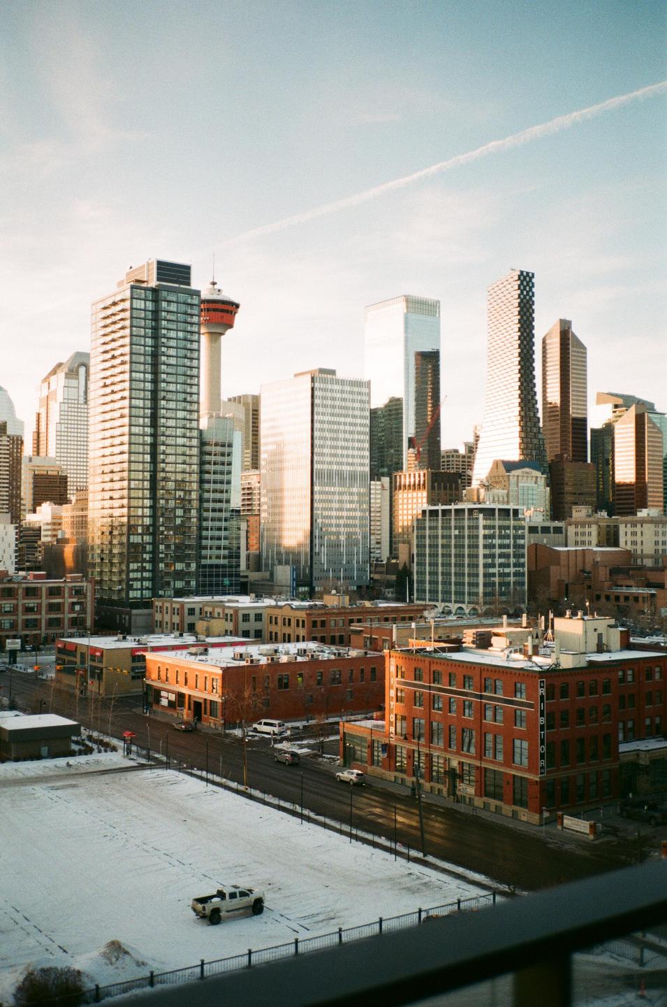 A cityscape of downtown Calgary shows tall buildings and a tower under a bright sky, with snow-covered ground and streets in the foreground.
