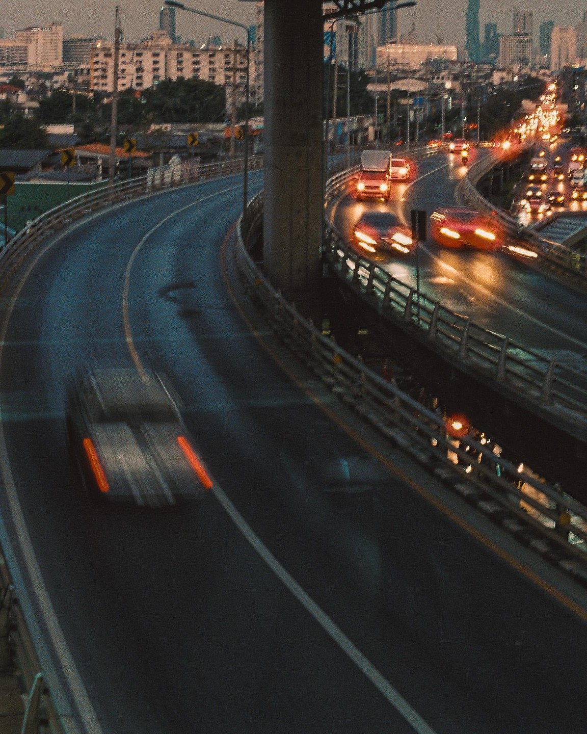 Cars drive on a multi-lane highway at dusk with city buildings in the background.