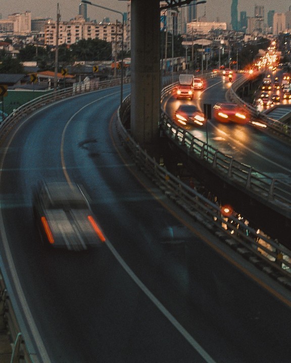 Cars drive on a multi-lane highway at dusk with city buildings in the background.