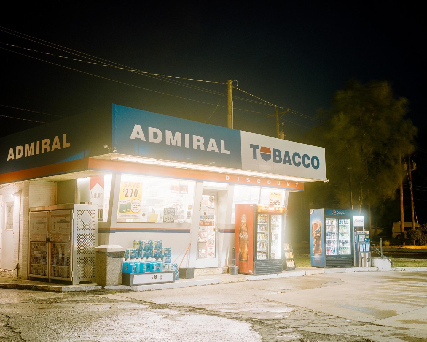 An Admiral Tobacco discount store is brightly lit at night, with several drink coolers and merchandise visible outside.