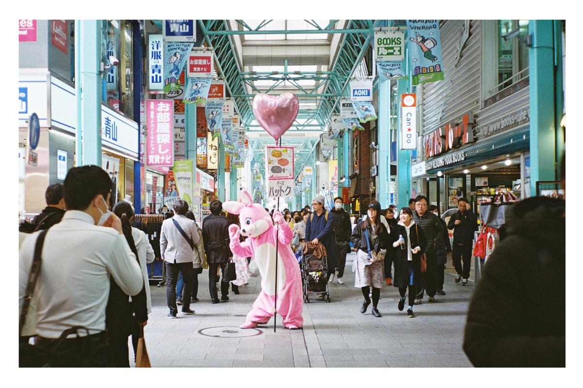A person in a pink rabbit costume holds a heart balloon in a busy Tokyo shopping street.