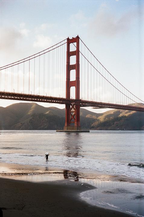 A person carries a surfboard into the water near the Golden Gate Bridge at golden hour.