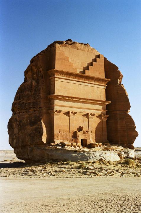 A large, ancient, carved rock structure stands in a desert landscape under a clear blue sky.