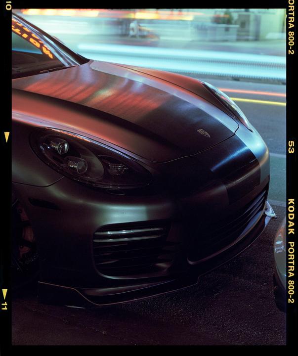 A dark vehicle is parked on a street at night, with reflections of neon lights on its hood.