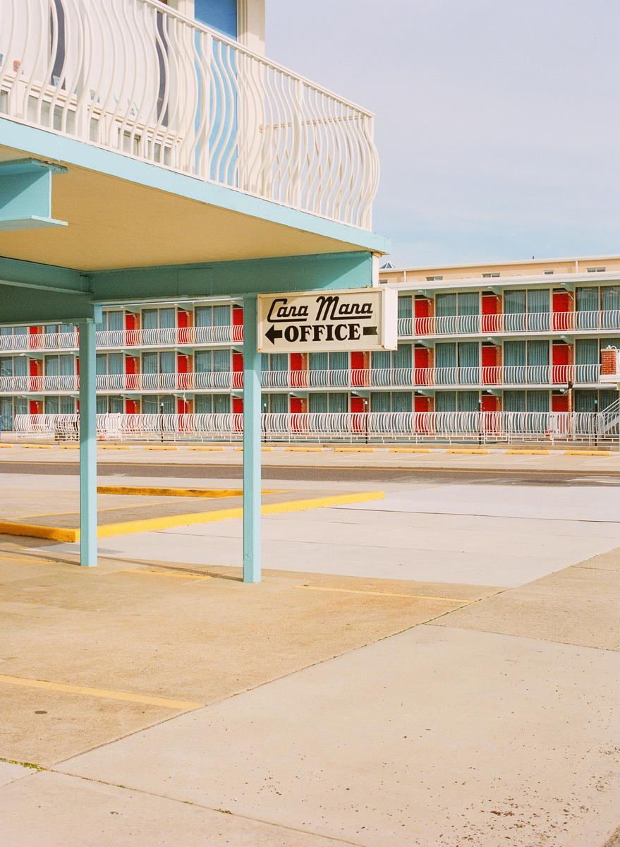 A sign for the Cara Mara office hangs under a teal awning in front of a motel with red doors and white railings.