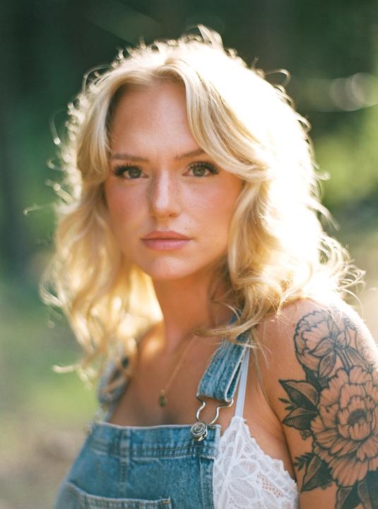 A woman with blonde hair and a floral tattoo on her shoulder looks directly at the viewer.