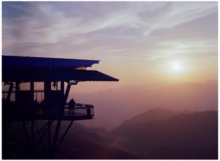 A building with a large deck overlooks a mountain range at sunset.