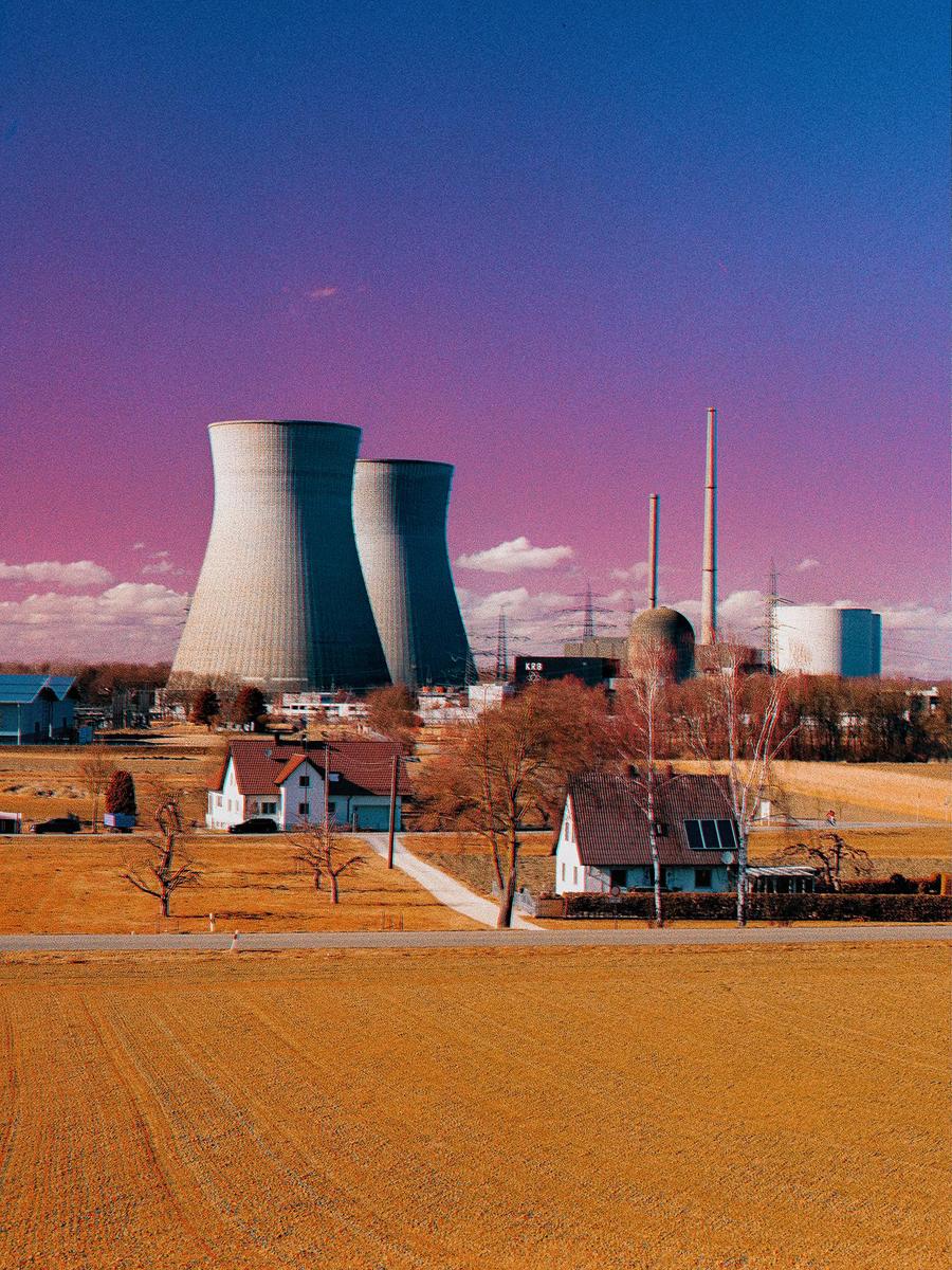 A nuclear power plant with two cooling towers stands behind houses and a field under a purple and blue sky.
