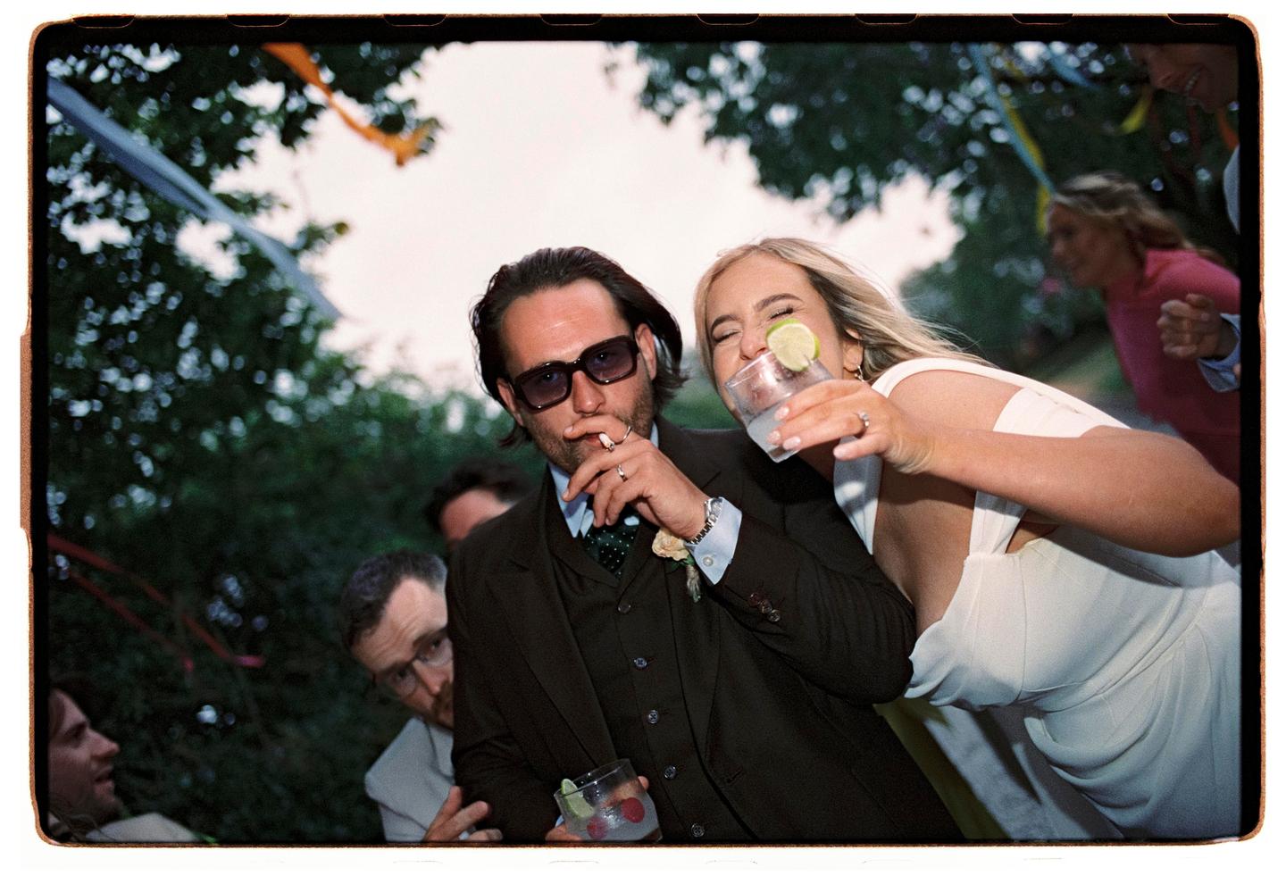 A man in sunglasses smokes a cigarette while a woman next to him laughs and holds a drink with a lime.