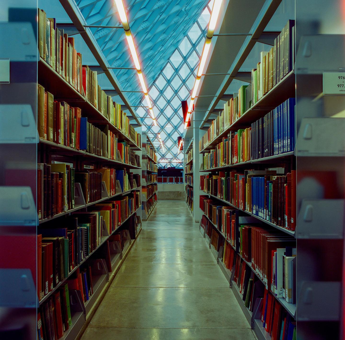 A long aisle in a library is lined with bookshelves and illuminated by a patterned skylight.