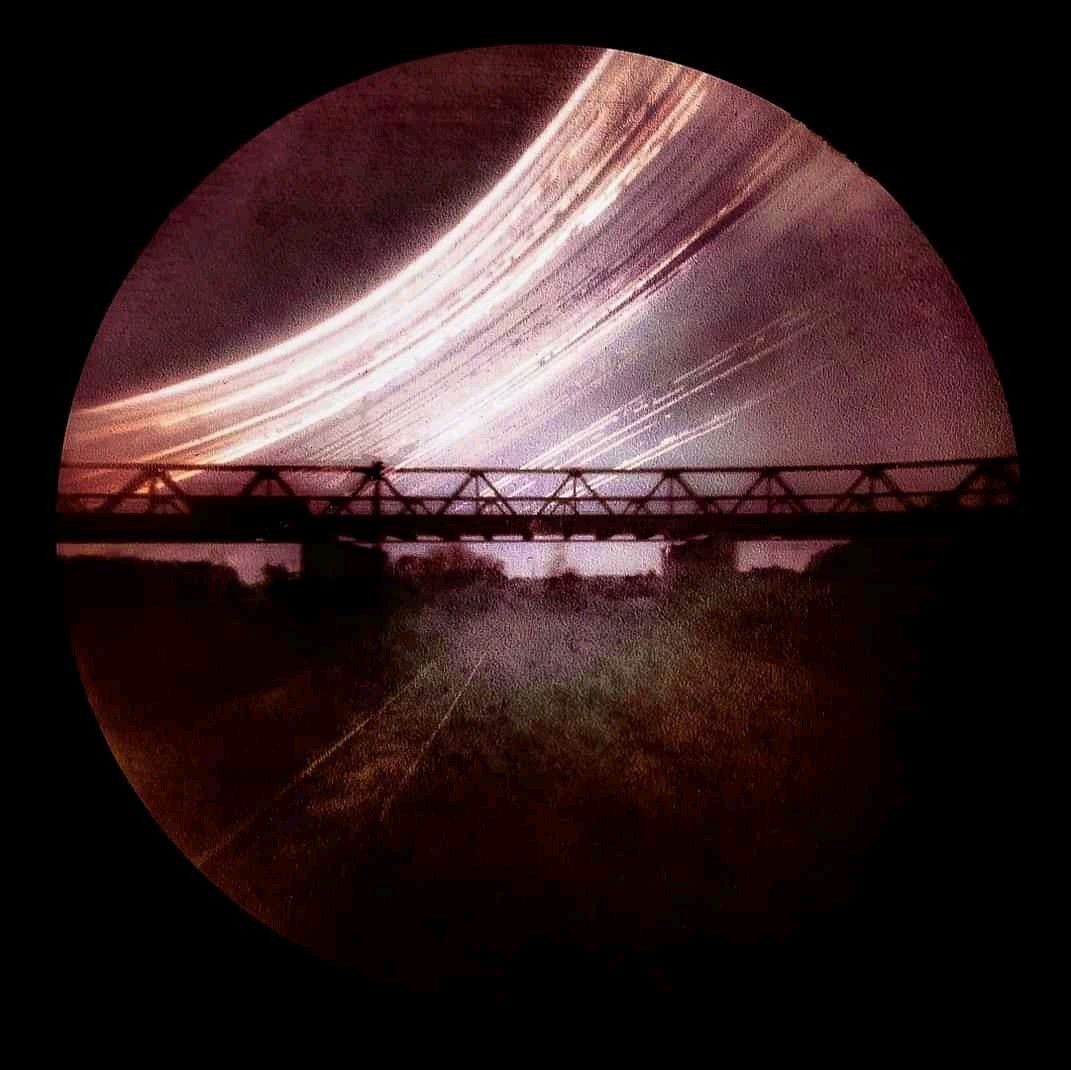 A long exposure captures the sun's path over a bridge and railway tracks.