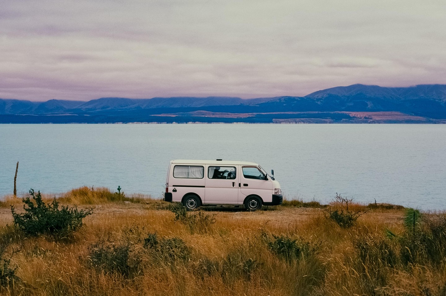 A white camper van is parked on a grassy hill overlooking a large lake with mountains in the distance under a cloudy sky.