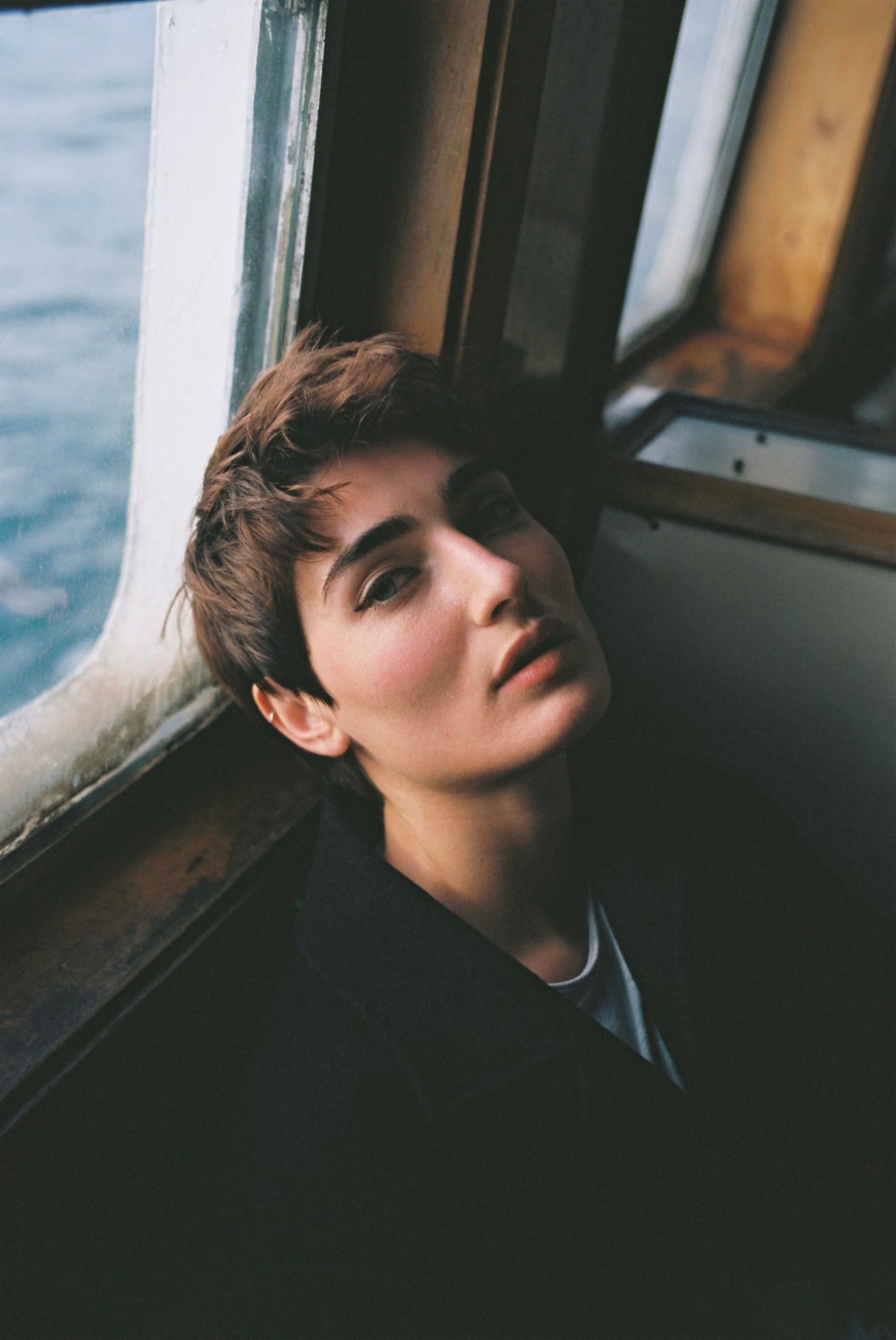 A woman with short hair looks up while sitting next to a window with water visible outside.