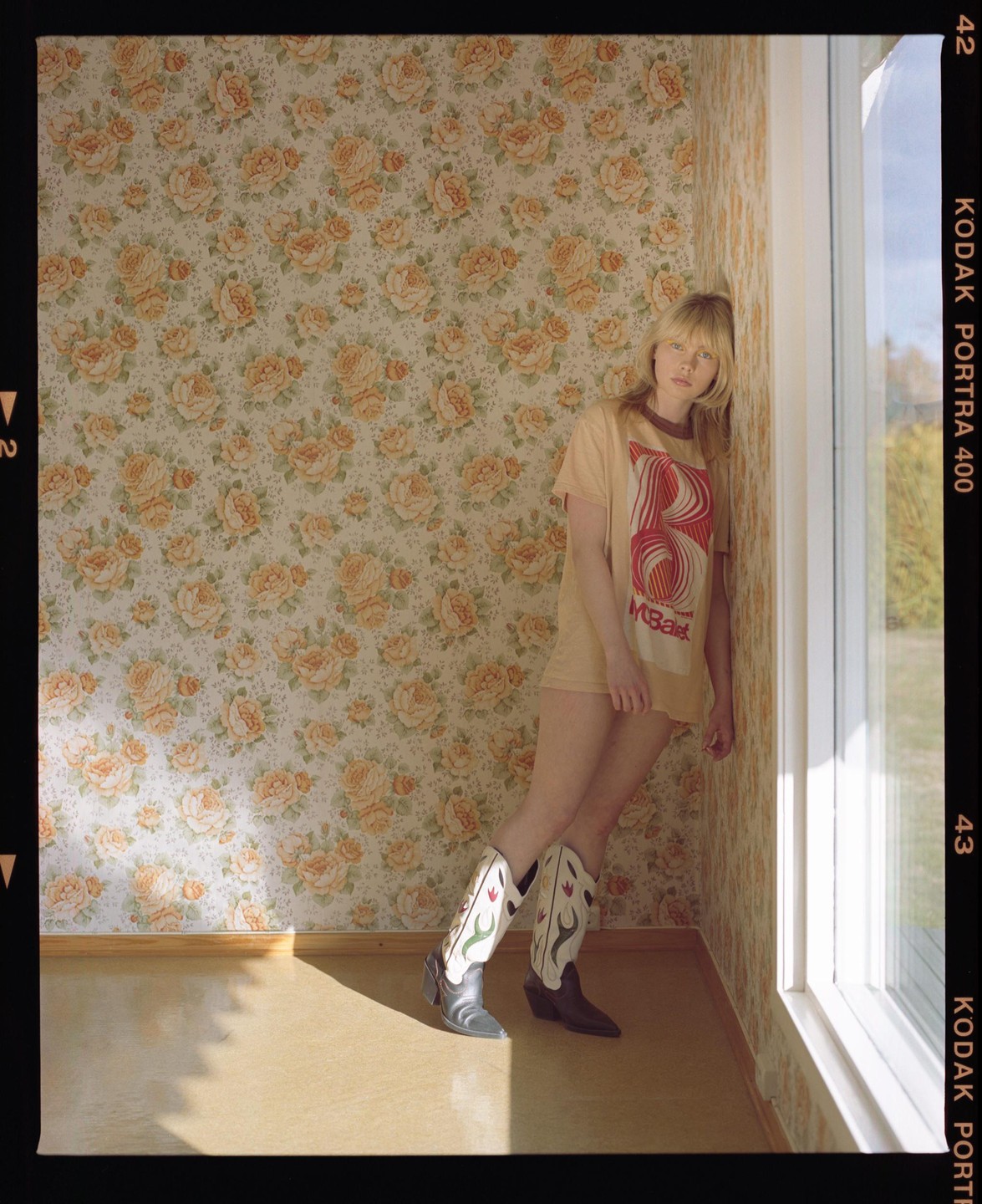 A woman wearing a t-shirt and cowboy boots leans against a floral wallpapered wall next to a window.