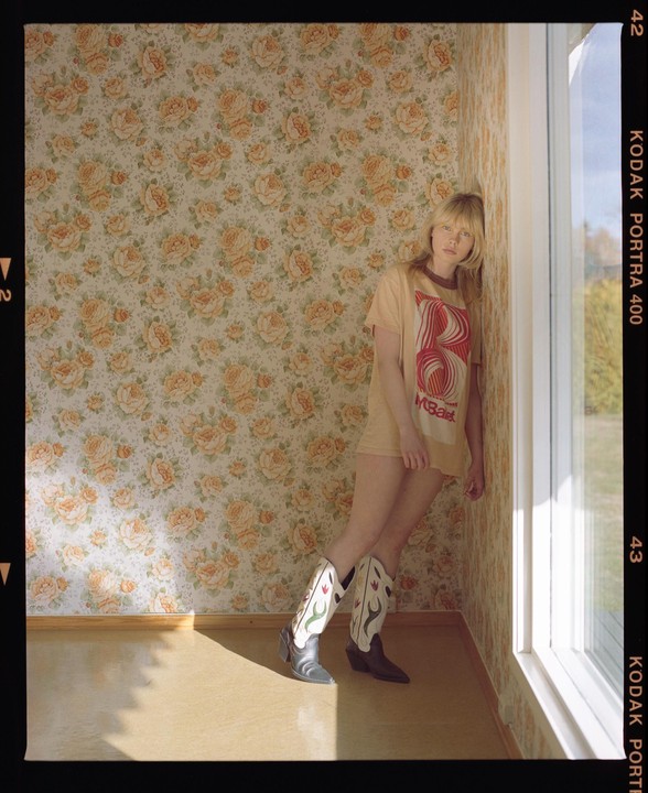 A woman wearing a t-shirt and cowboy boots leans against a floral wallpapered wall next to a window.
