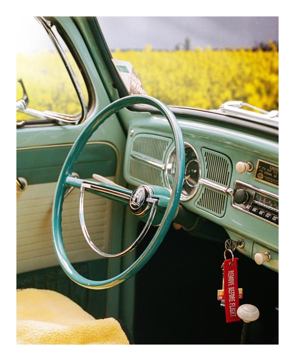 The interior of a vintage car with a teal steering wheel is parked in front of a field of yellow flowers.