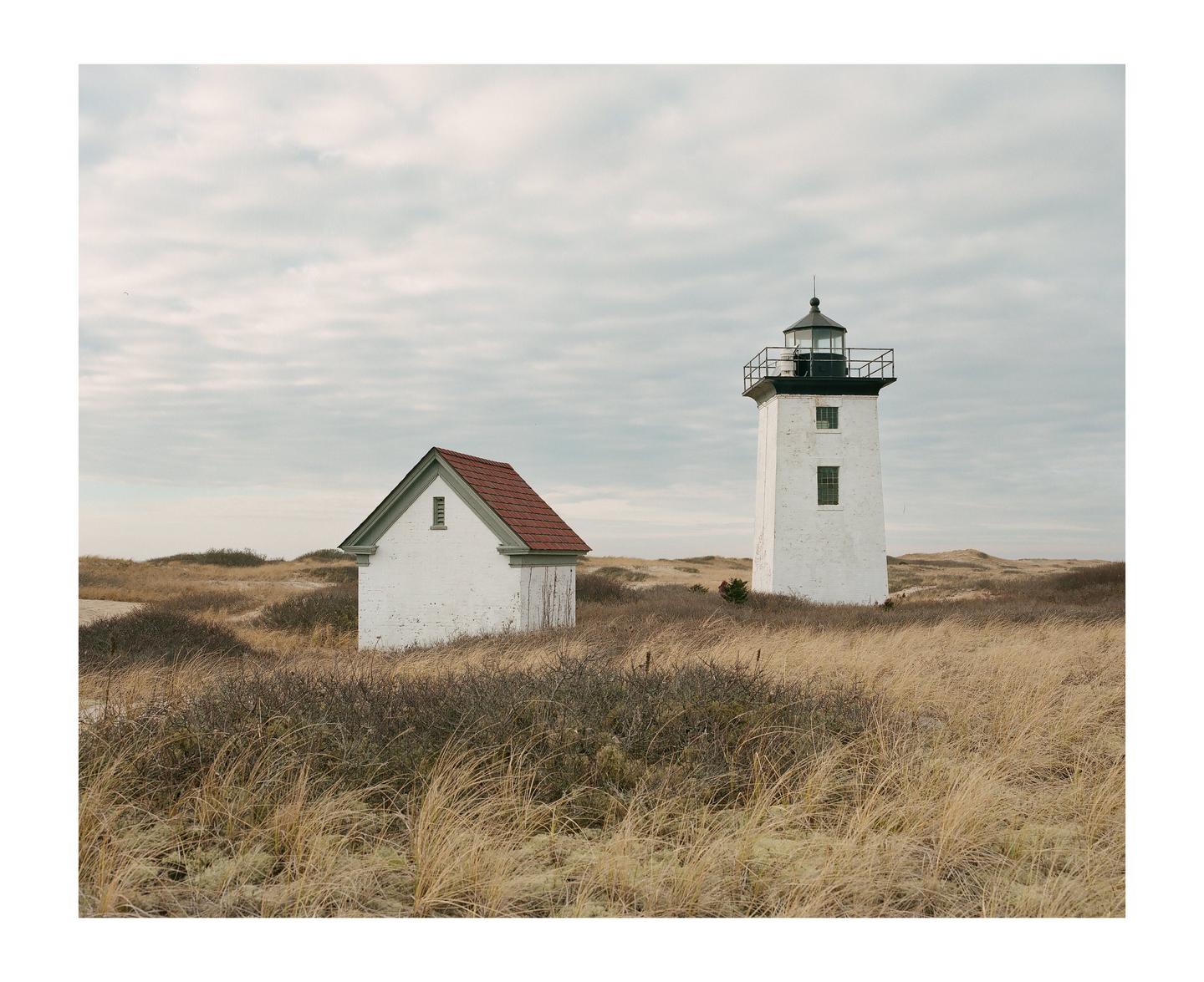A white lighthouse and a small white building with a red roof stand in a field of tall, dry grass under a cloudy sky.
