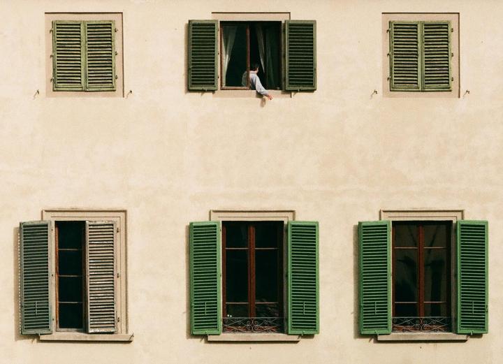 A person leans out of an open window with green shutters on a building facade.