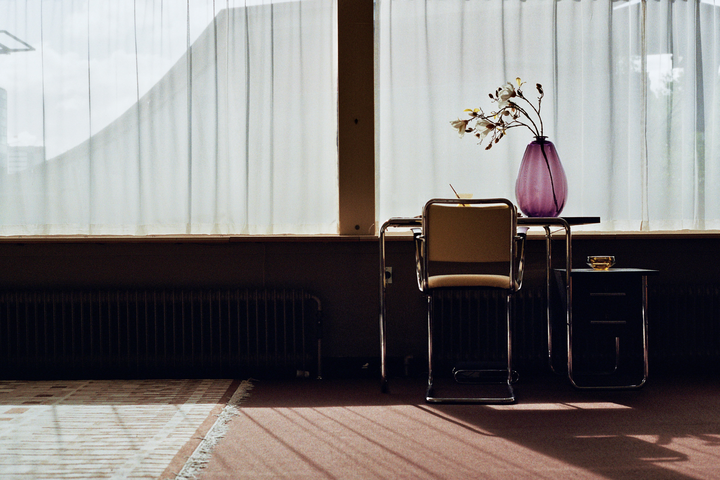 A purple vase with flowers sits on a table next to a chair in front of a window with sheer white curtains.