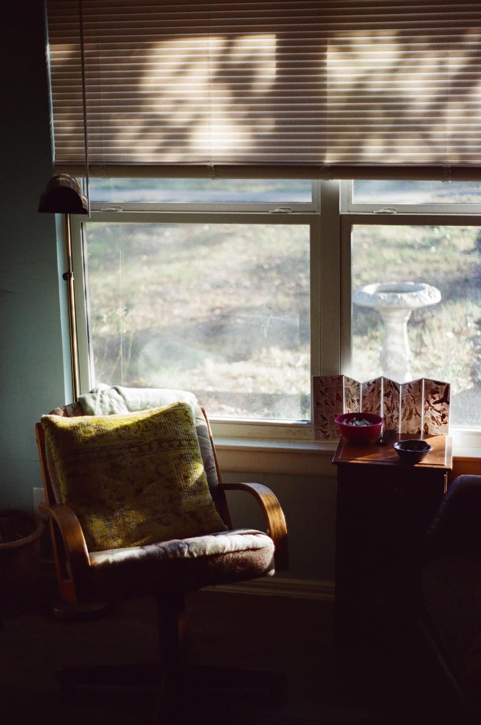 Sunlight streams through a window with blinds, illuminating a chair with a pillow and a small table with bowls and a decorative screen.