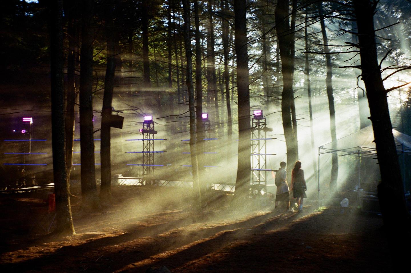Two people stand in a hazy forest with sunbeams shining through the trees and stage equipment in the background.