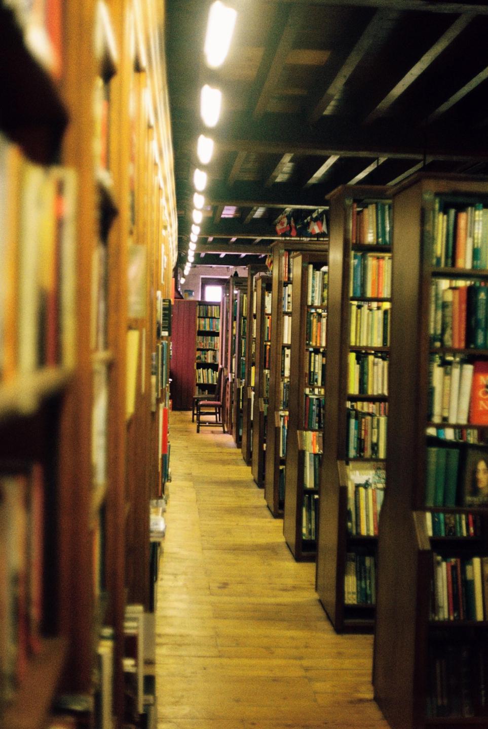 A long, narrow aisle in a library is lined with tall wooden bookshelves filled with books, with a chair visible at the far end.