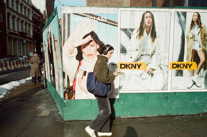A woman wearing headphones walks past a large billboard on a city street with another person in the distance.