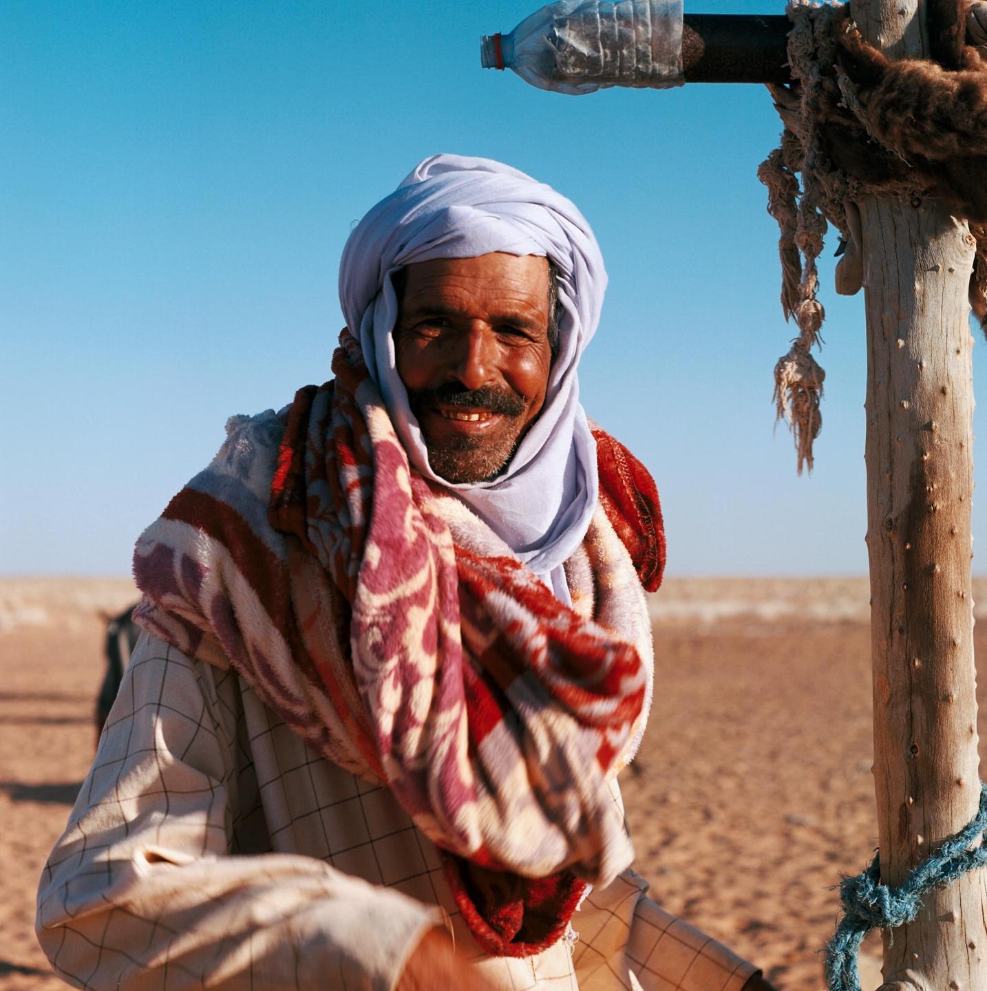 A man wearing a turban and a patterned blanket smiles at the camera in a desert landscape.