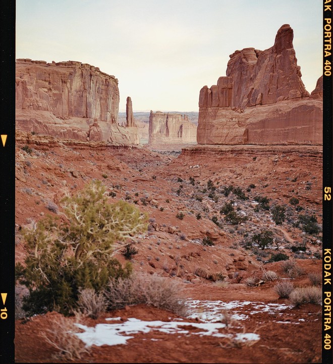 A vast desert landscape features towering red rock formations under a pale sky, with sparse vegetation and patches of snow on the ground.