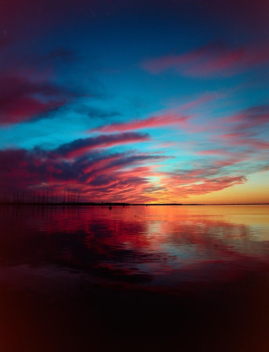 A vibrant sunset over a body of water reflects colorful clouds, with a distant shoreline and boat masts visible.