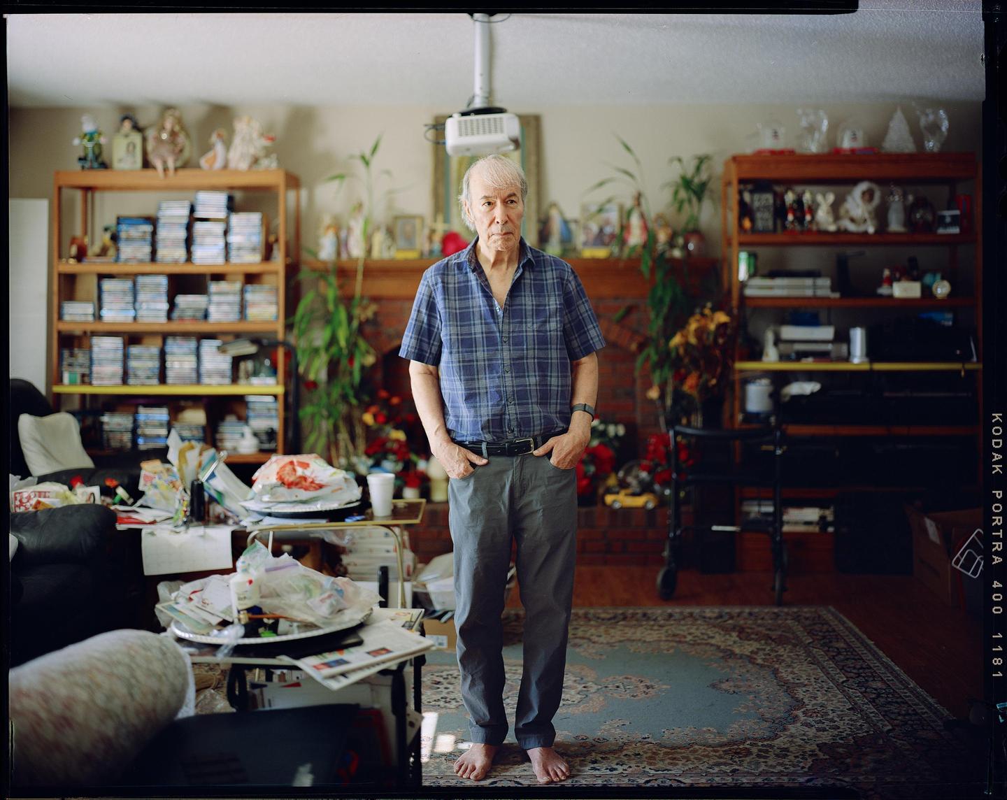 A man stands barefoot in a cluttered living room with shelves full of items and a patterned rug.