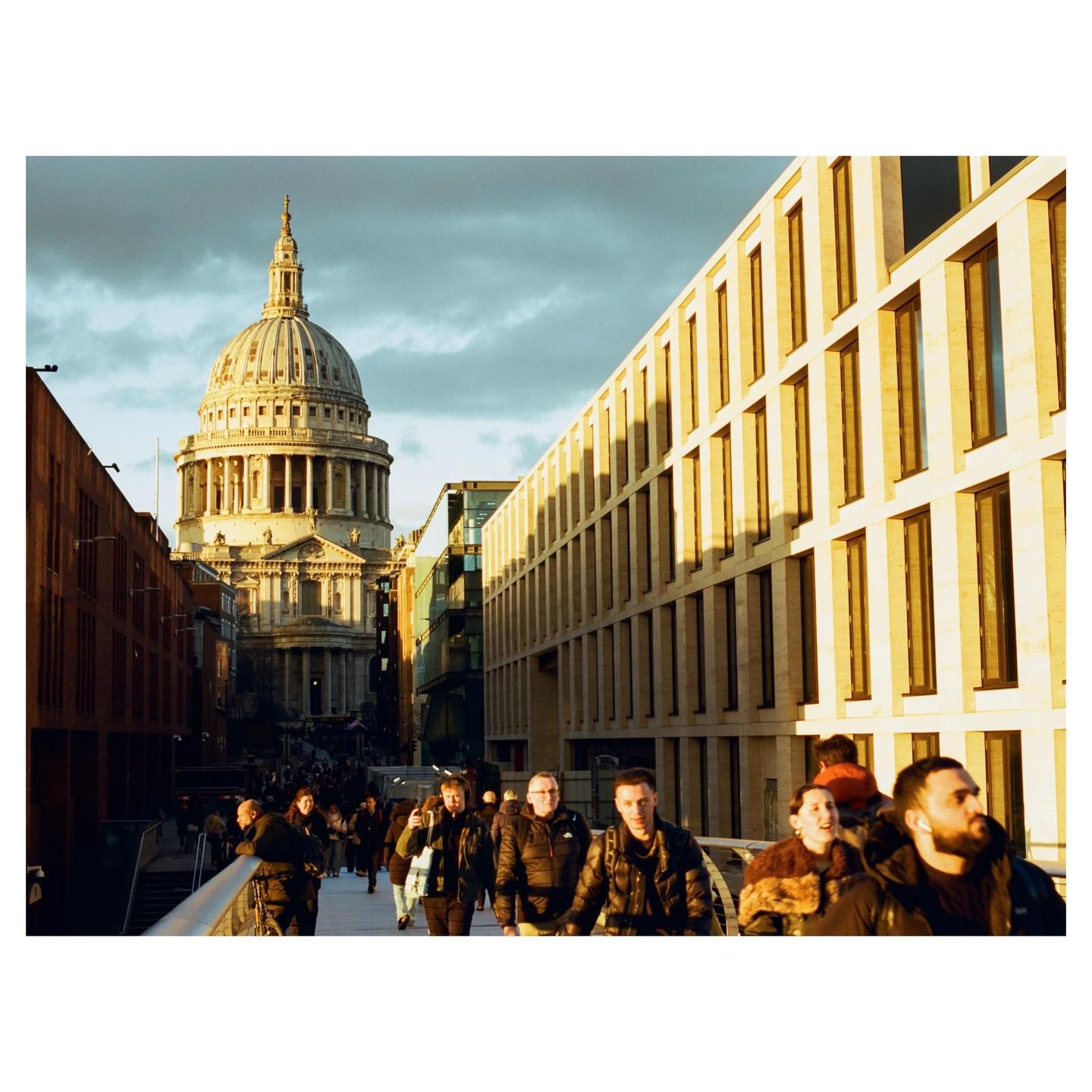 A crowd of people walk on a bridge with a large domed building in the background.