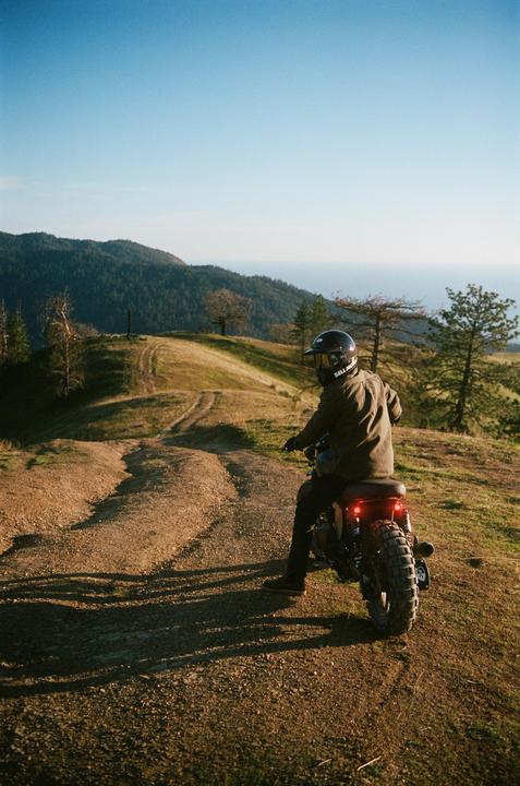 A person on a motorcycle is parked on a dirt road overlooking a mountainous landscape and the ocean.