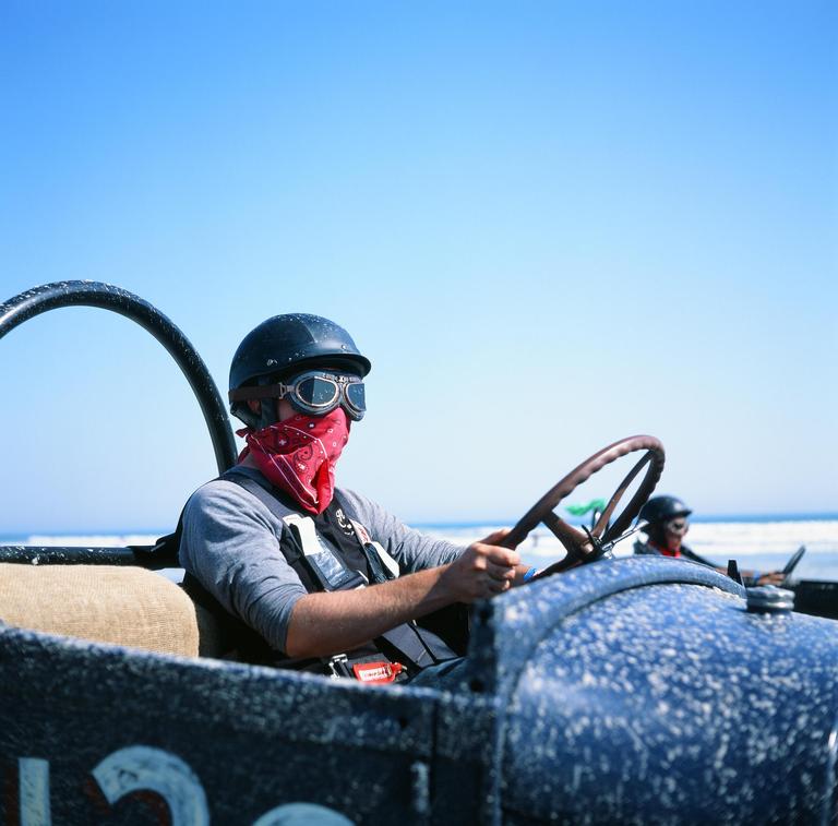 A man in a helmet and goggles sits in a vintage race car on a beach.