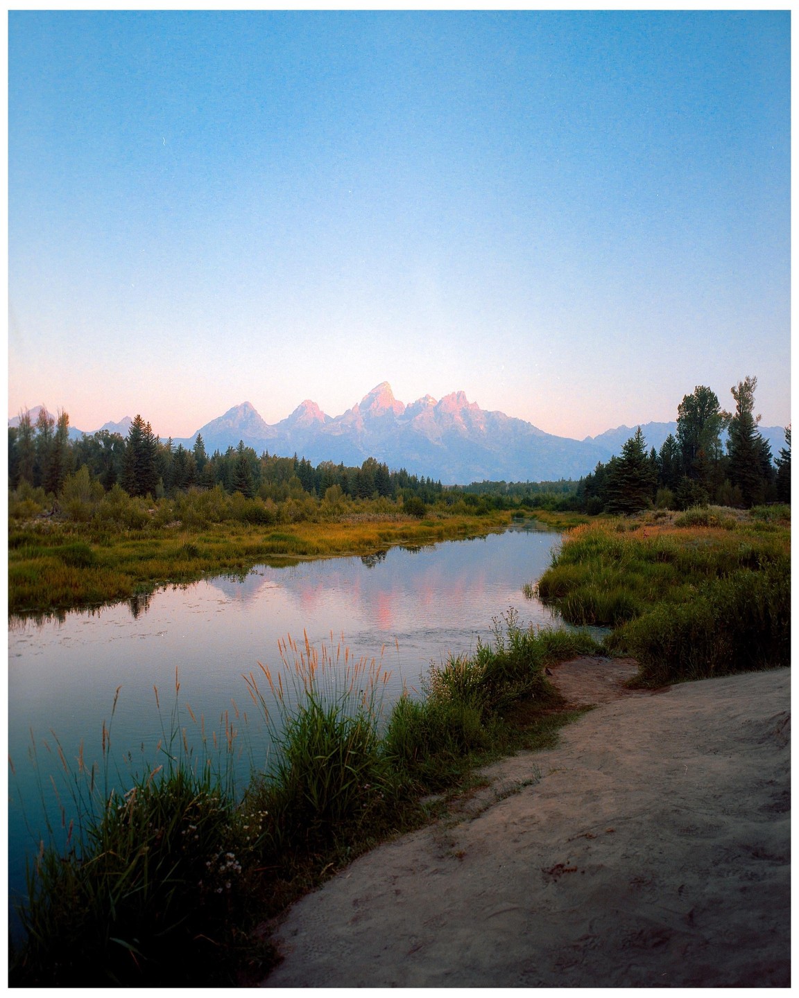 A river flows through a valley with mountains in the background under a clear sky.