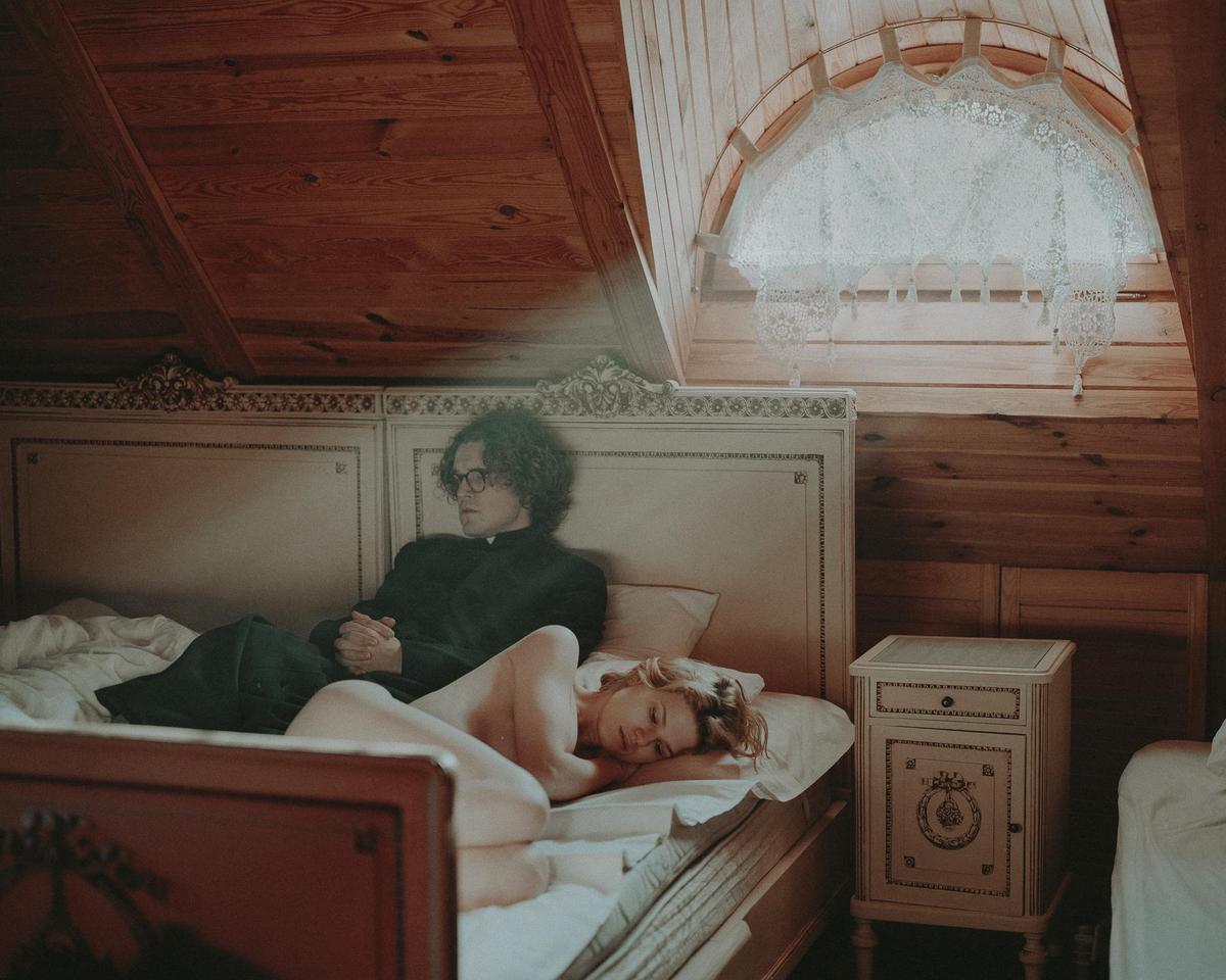 A man in a dark suit sits on a bed next to a sleeping woman in a room with wood paneling and a lace-covered window.