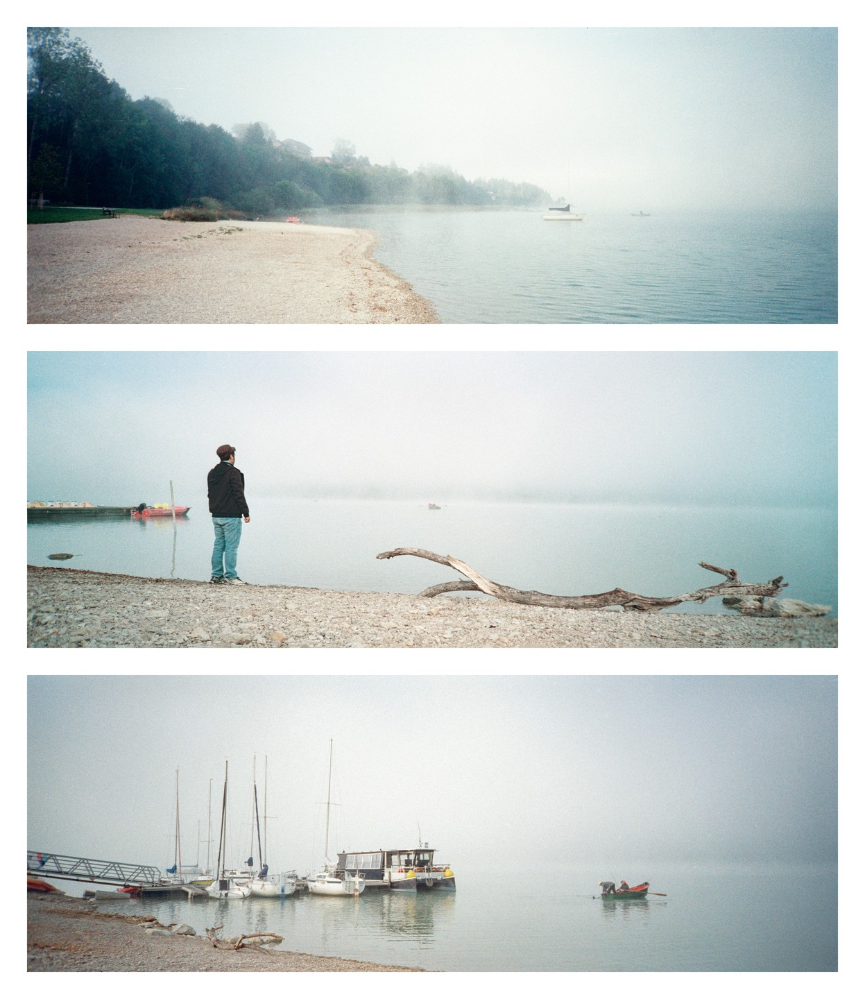 A man stands on a rocky beach looking out at a foggy lake with boats and a forest in the distance.