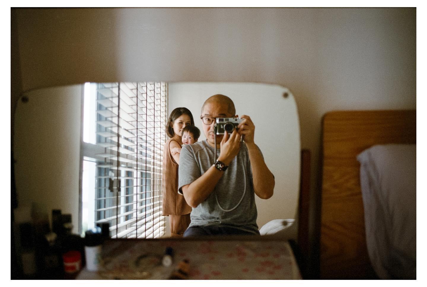 A man takes a self-portrait in a mirror, with a woman and child visible behind him.