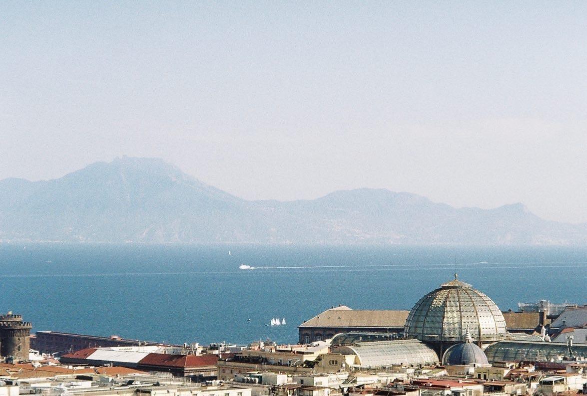 A city with a large domed building in the foreground overlooks a body of water with mountains in the distance.