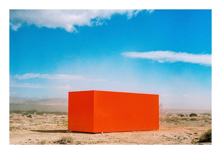 A large orange rectangular box sits in a desert landscape under a blue sky with clouds.