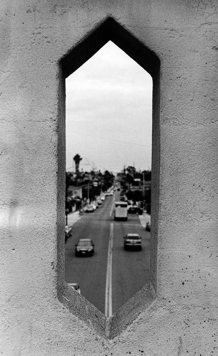A street with cars and a bus is visible through a narrow, pointed opening in a concrete wall.