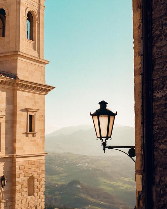 A street lamp is mounted on a stone wall with a view of mountains and a valley in the background.