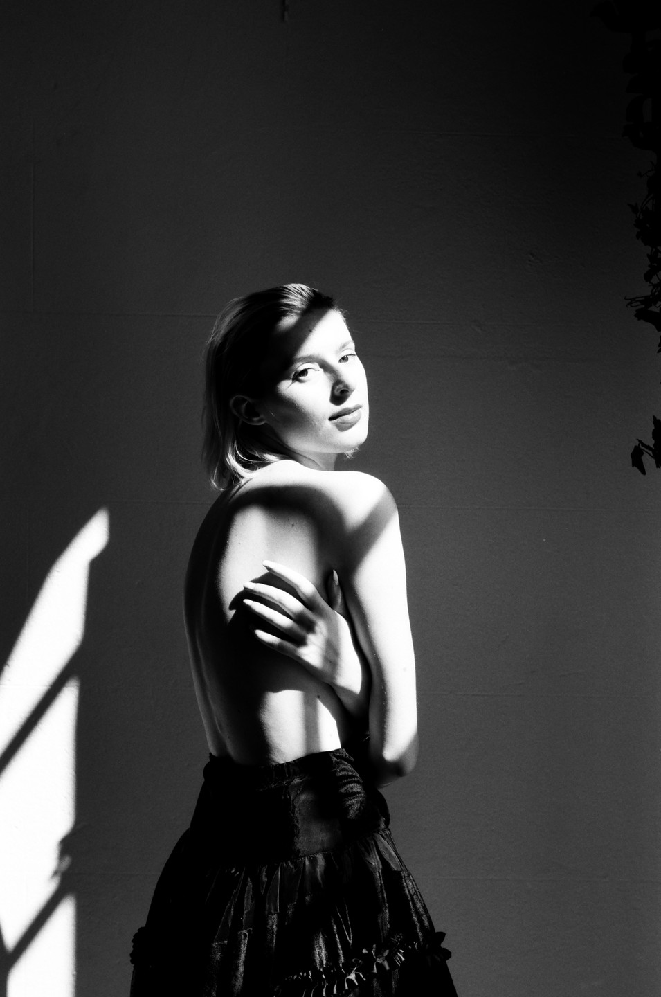 A woman with bare shoulders and a ruffled skirt looks over her shoulder, with a strong shadow cast across her face and body.