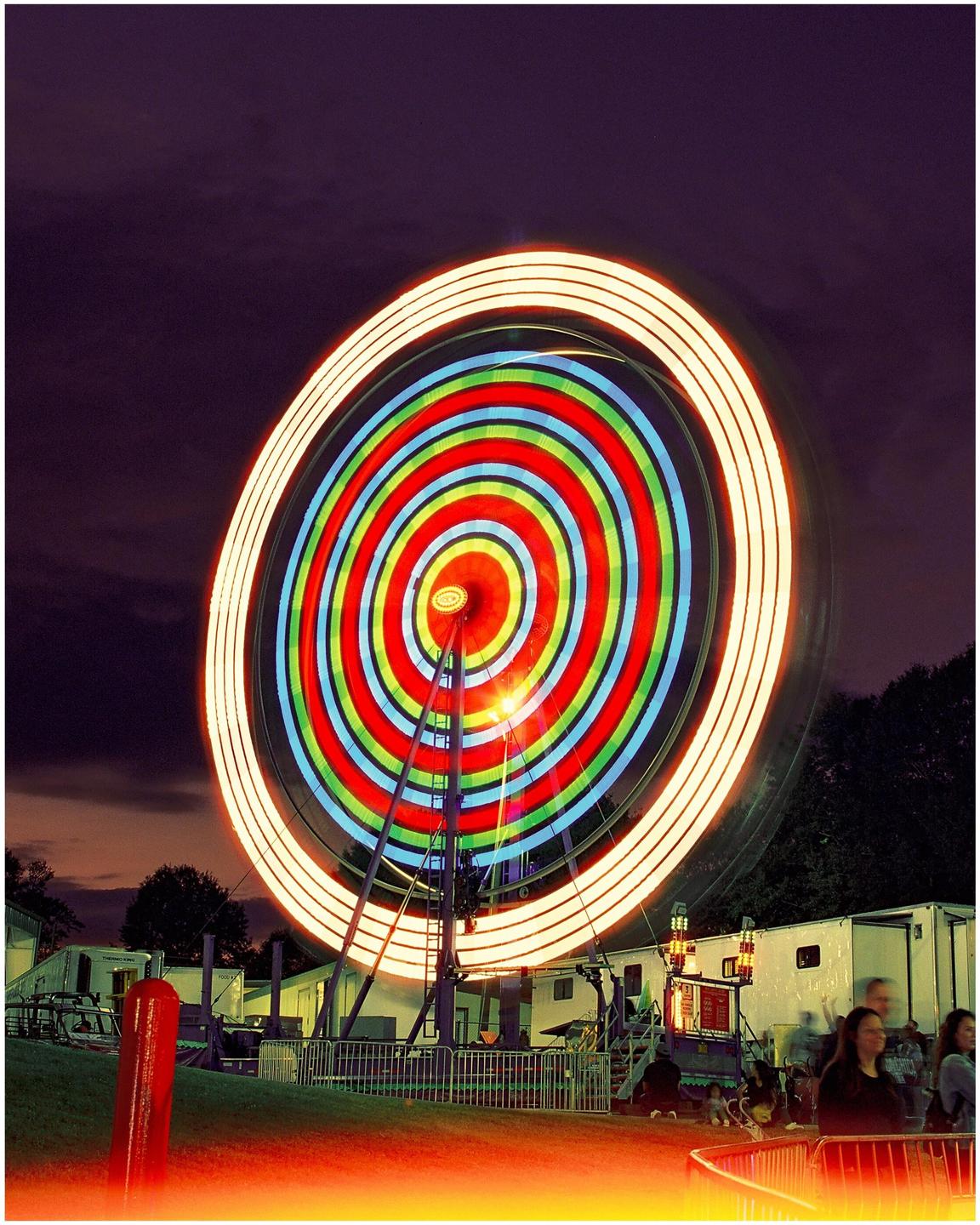 A long exposure photograph captures the colorful, spinning lights of a carnival ride against a dark night sky.