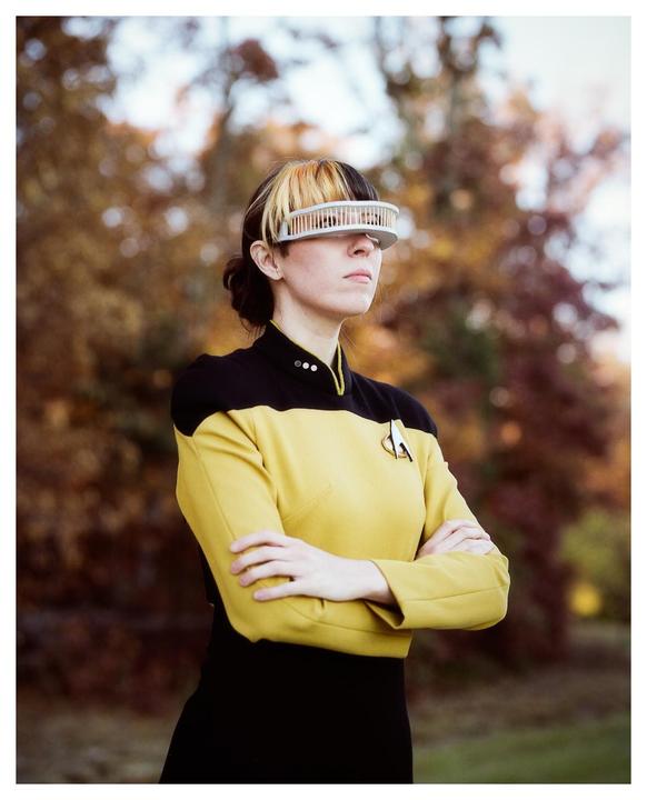 A woman in a yellow and black uniform with a futuristic visor stands with her arms crossed outdoors.