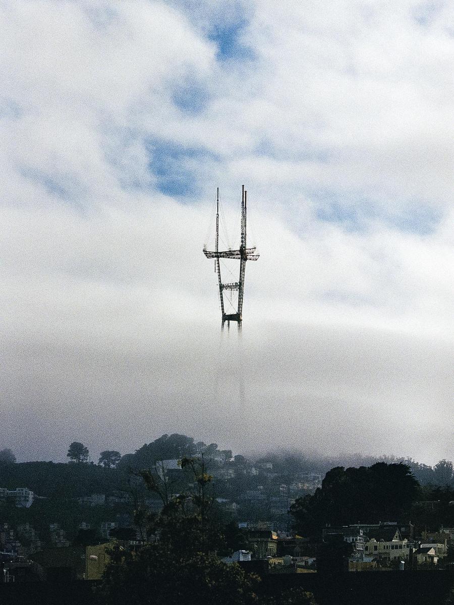 A tall radio tower rises above a layer of fog, with a city and hills visible below.