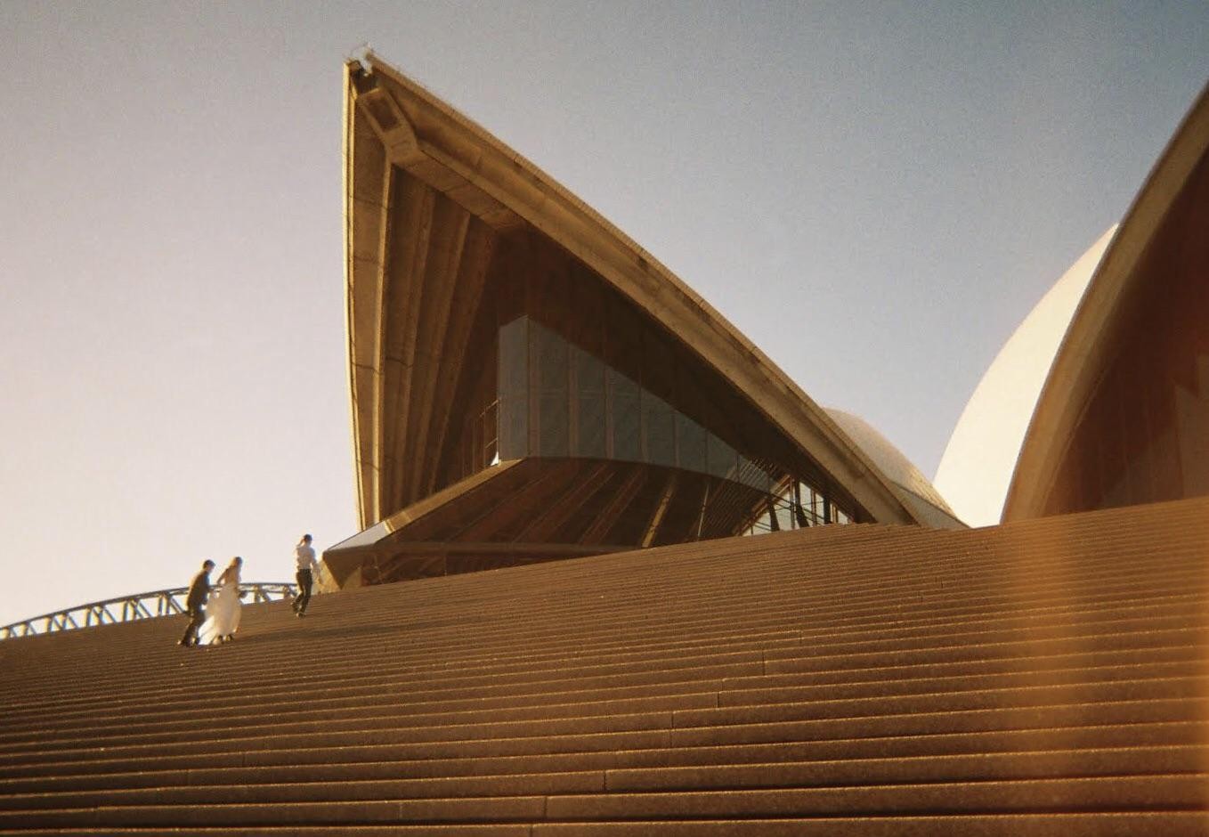 Three people ascend a wide staircase towards the Sydney Opera House.